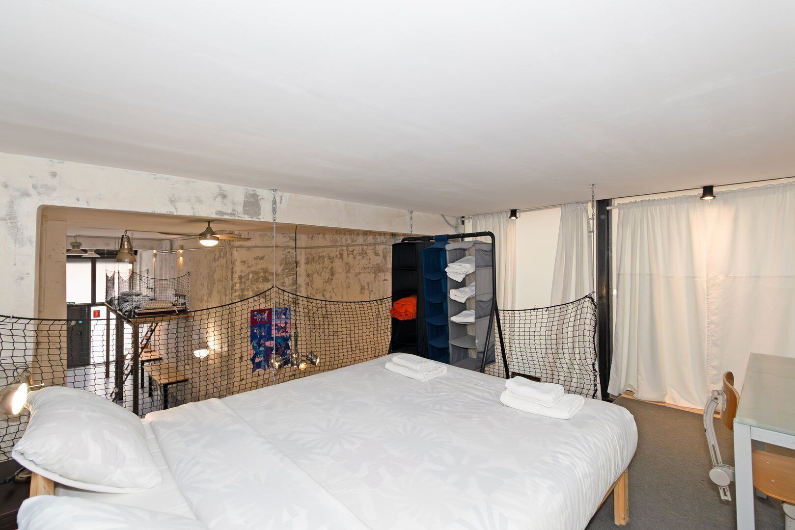Habitación de hotel con una cama grande, un escritorio y una silla. Se ve una ventana con cortinas, una puerta y una pared con un diseño texturizado.