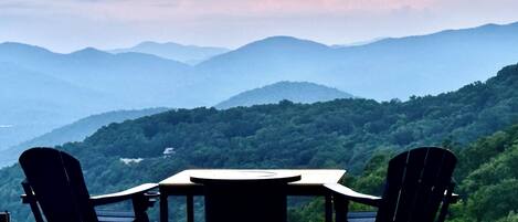 A wooden deck with two Adirondack chairs and a table, overlooking a mountainous landscape.
