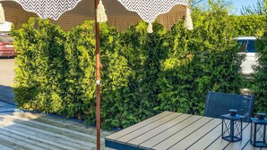Eine Holzterrasse mit Tisch, Stühlen und einem Sonnenschirm, im Hintergrund ein Heckenstreifen.