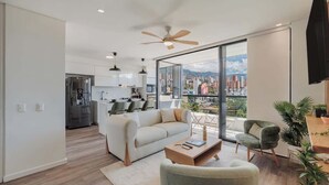 A modern living room with a sofa, coffee table, and armchairs, a dining area with a table and chairs, a kitchen with white cabinets, and a large sliding glass door leading to a balcony with a city view. - lEPl Nice Apt w/Pool, Good Zone, Bbq & Balcony (Medellín)