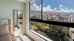 A room with a large window offering a cityscape view, a bed, and a framed mirror. - lEPl Nice Apt w/Pool, Good Zone, Bbq & Balcony (Medellín)