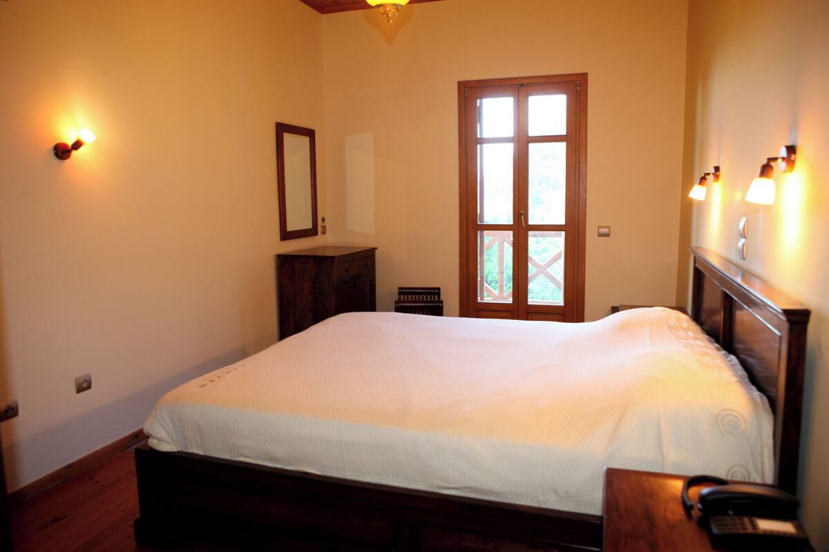 Un dormitorio con cama, cómoda de madera, espejo, puerta con ventanales e iluminación en la pared.
