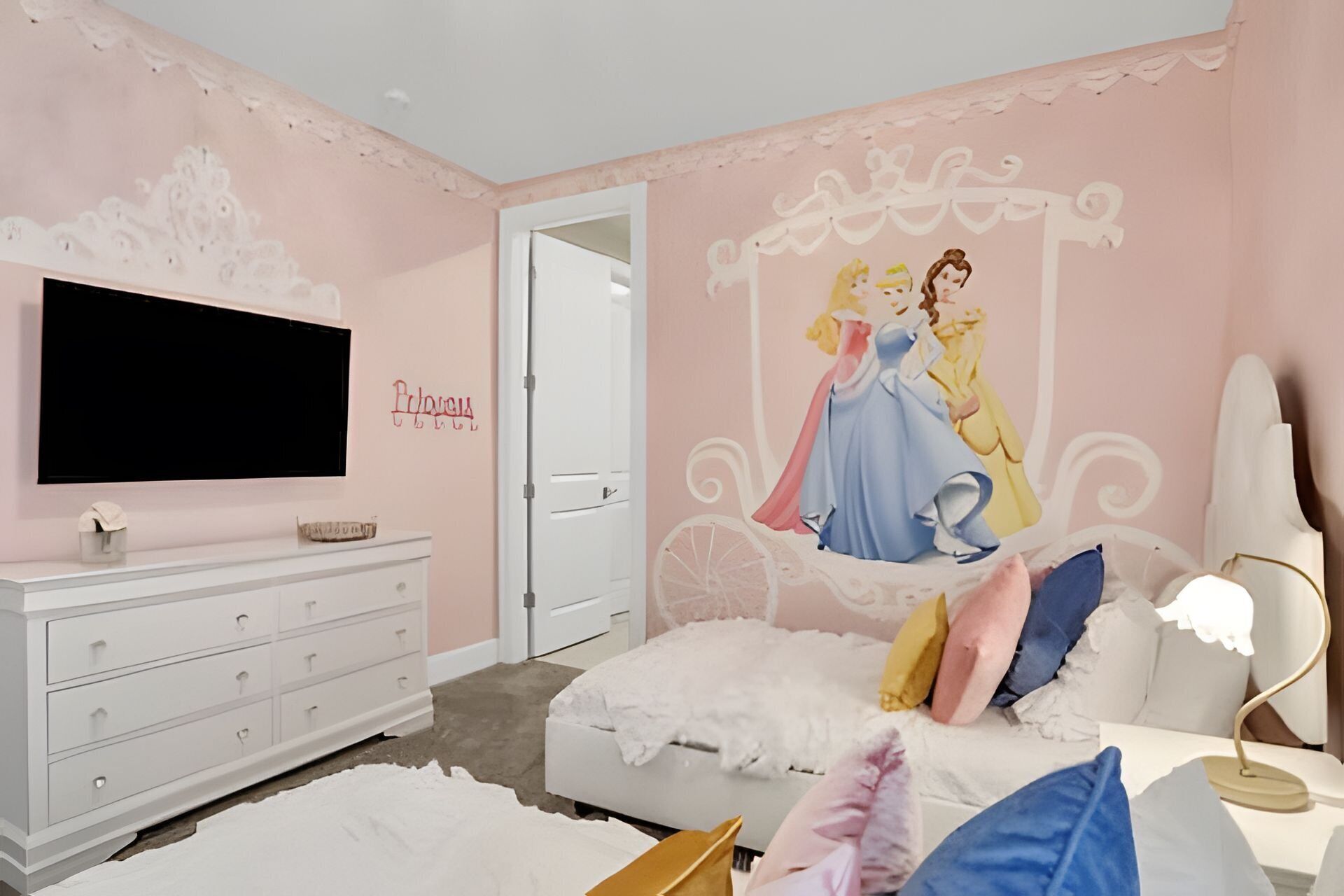 A bedroom with a pink wall, a white dresser, a bed with colorful pillows, a white nightstand, a lamp, and a Disney princess mural.