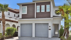 A modern two-story house with two garage doors, a flat roof, and a well-maintained garden with palm trees. - Reunion Luxury Villa w/ Lake View & Game Room (Kissimmee)