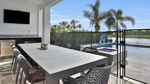 A modern outdoor dining area with a large table and chairs, a TV mounted on the wall, and a view of a swimming pool and palm trees. - Reunion Luxury Villa w/ Lake View & Game Room (Kissimmee)