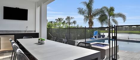 Ein moderner Essbereich im Freien mit einem großen Tisch und Stühlen, einem an der Wand montierten Fernseher und Blick auf einen Swimmingpool und Palmen.