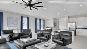 A modern living room with black leather sofas, a ceiling fan, and a kitchen in the background. - Reunion Luxury Villa w/ Lake View & Game Room (Kissimmee)