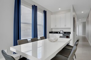 A modern dining room with a white table and chairs, a kitchen area with white cabinets, and blue curtains on the windows. - Reunion Luxury Villa w/ Lake View & Game Room (Kissimmee)