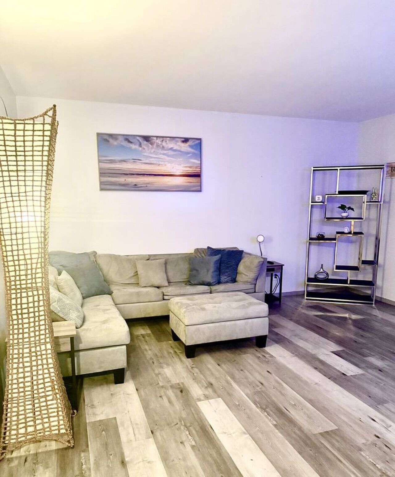 A living room with a sectional sofa, a wooden floor, a wall-mounted shelf, and a painting.
