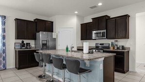 A modern kitchen with dark wood cabinets, a stainless steel refrigerator, a microwave, a stove, and a granite countertop. There are three bar stools and a white door. - 5BR Villa w/ Pool + Resort Access – Solterra (Davenport)