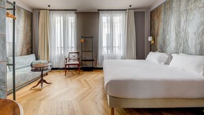 A hotel room with a large bed, a sofa, a small table, and a chair. There are curtains on the windows, and a lamp on the wall. - Hôtel Maât Batignolles (Paris)