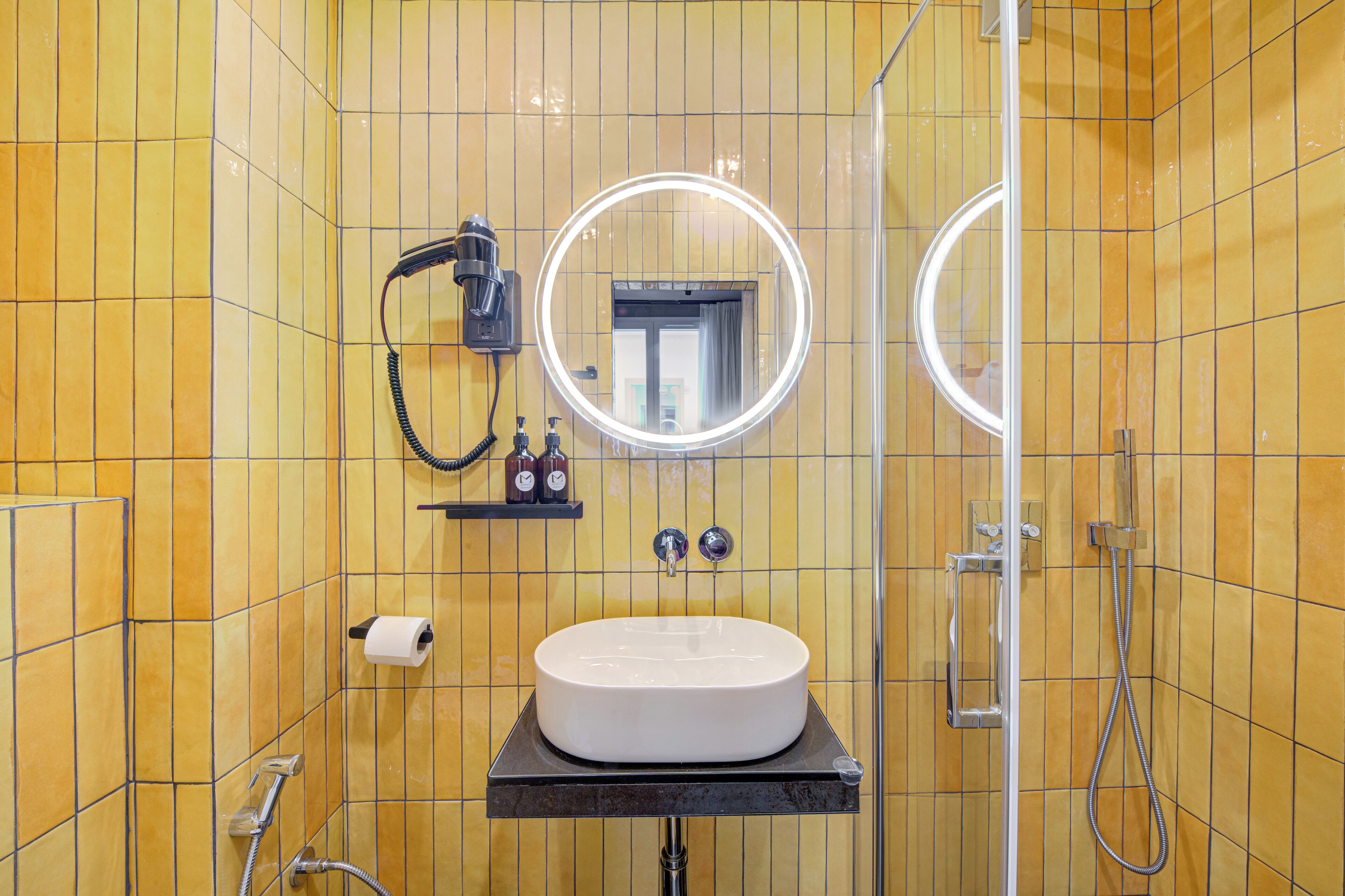 Un bagno con pareti piastrellate di giallo, un lavandino bianco, uno specchio con illuminazione e una zona doccia.