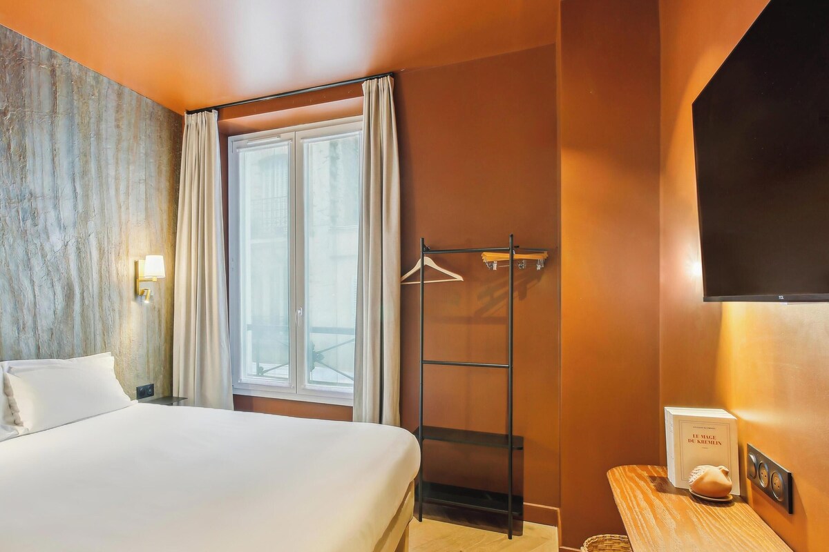 A hotel room with a large bed, a window with curtains, a wall-mounted TV, and a wooden side table with a book and a decorative item.