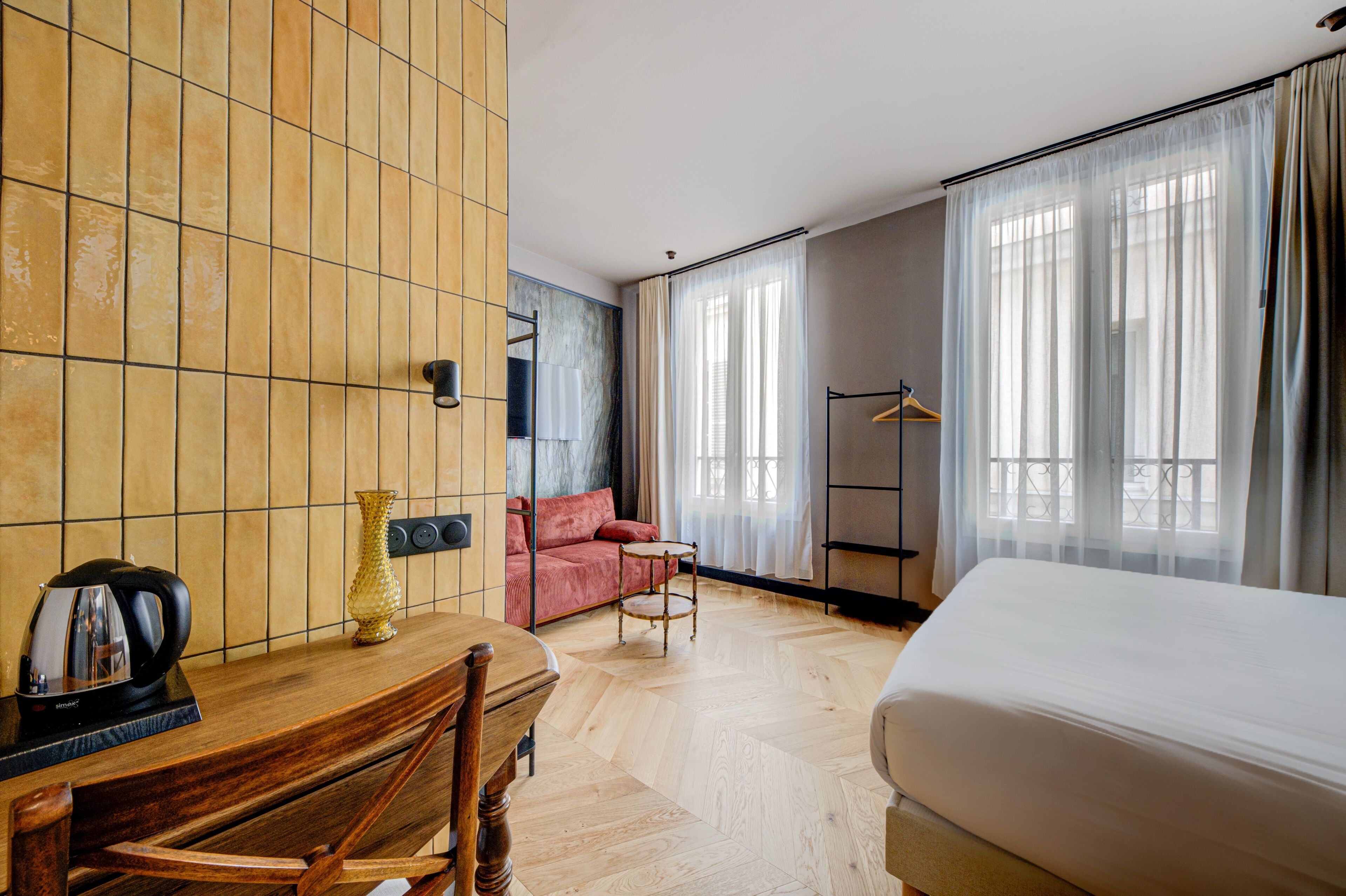 A hotel room with a wooden floor, a bed, a desk with a kettle, a chair, a sofa, and a large window with curtains.