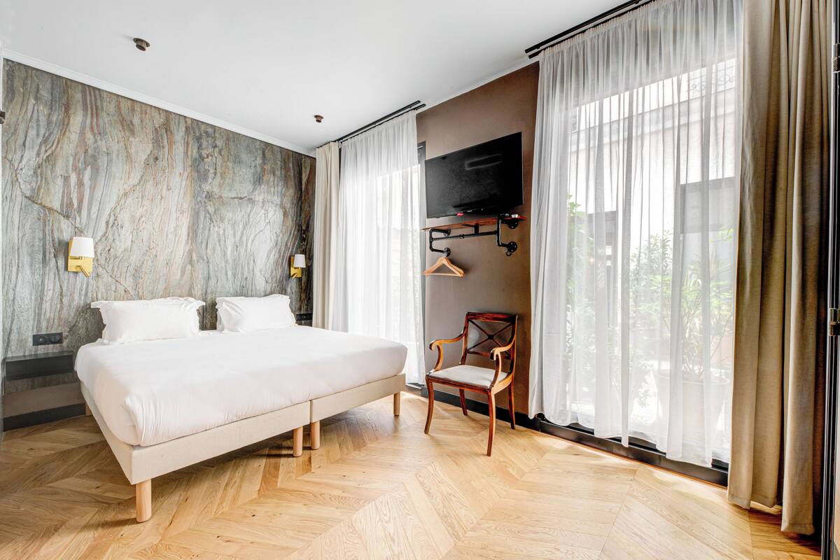 A modern hotel room with a large bed, a wooden floor, a TV mounted on the wall, and a chair.