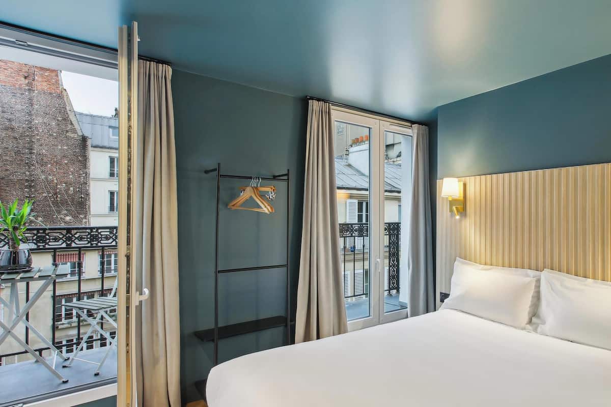 A modern hotel room with a large bed, a view of the city through a window, a balcony with a plant, and a hanging coat rack.