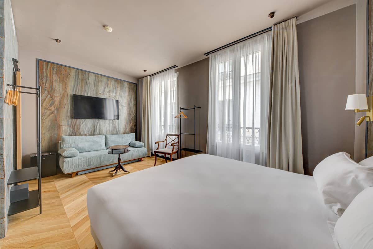 A modern hotel room with a large bed, a sofa, a small table, and a TV mounted on a marble wall.