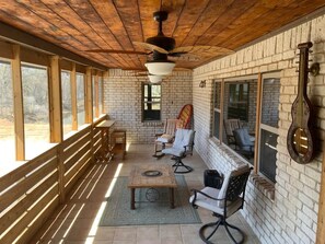 Un espacio de estar al aire libre cubierto con bancos de madera, una mesa y sillas, con paredes de ladrillo y ventanas grandes.