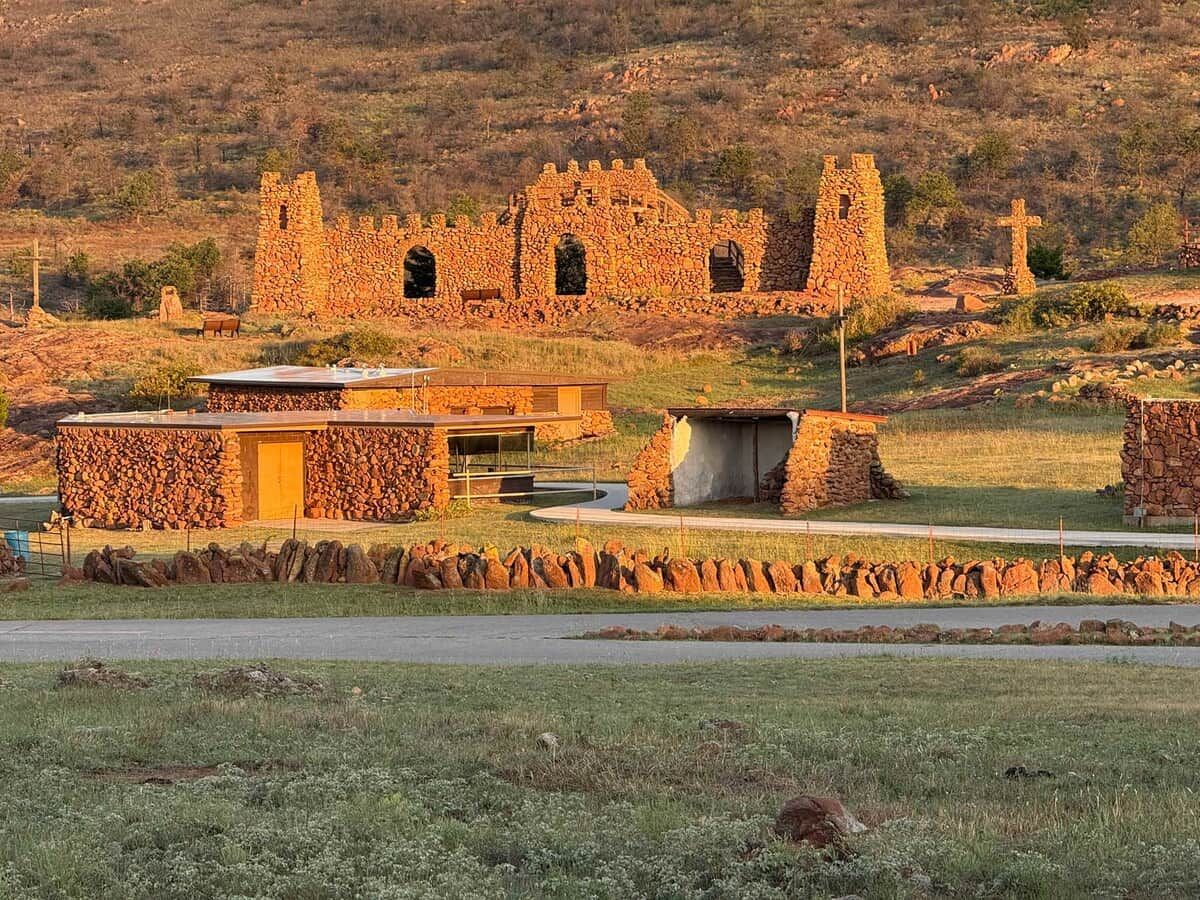Un edificio de piedra con una estructura moderna al frente, rodeado de un muro de piedra y un área con césped.