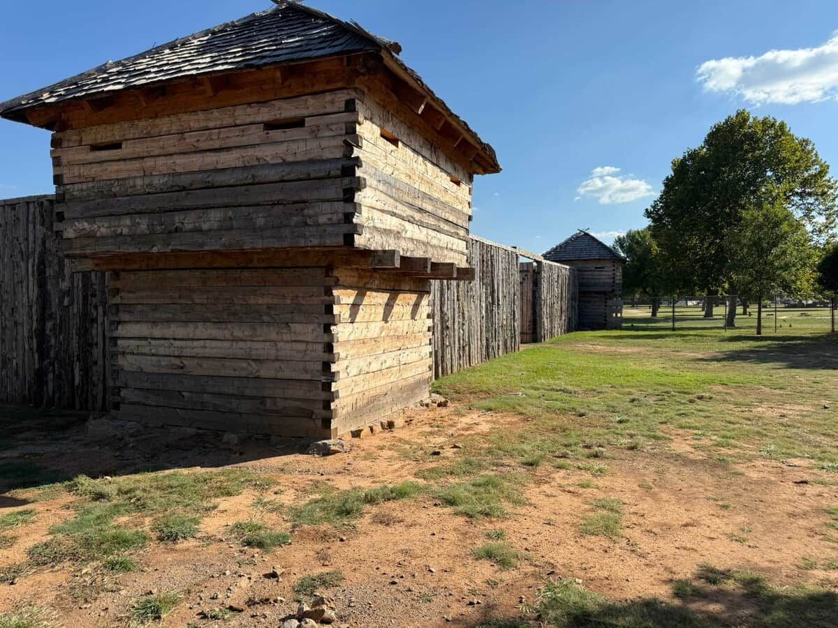 A rustic wooden structure with a sloped roof, surrounded by a fenced area with sparse grass and a few trees.