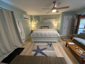 A bedroom with a bed, bedside lamps, a ceiling fan, a star-patterned rug, a wooden coffee table, and a view of the outdoors through a door.