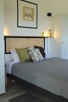 Una cama bien hecha con una colcha gris y varios almohadones, un cabecero tejido y un cuadro enmarcado sobre la cama.
