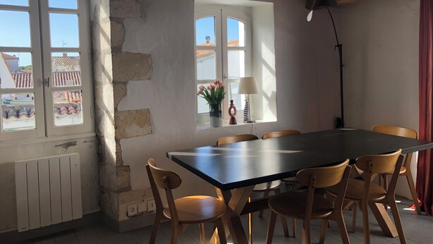 Een eetgedeelte met een grote tafel en zes stoelen, een raam met uitzicht op gebouwen, een radiator en een lamp.