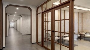 Un pasillo moderno de hotel con puertas de vidrio enmarcada en madera, un largo pasillo y un área de descanso con mesas y sillas.