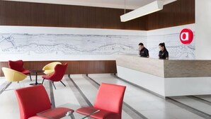 A modern reception area with a large desk, two receptionists, and red and yellow chairs. - Amaris Hotel Pettarani - Makassar (Panakkukang)