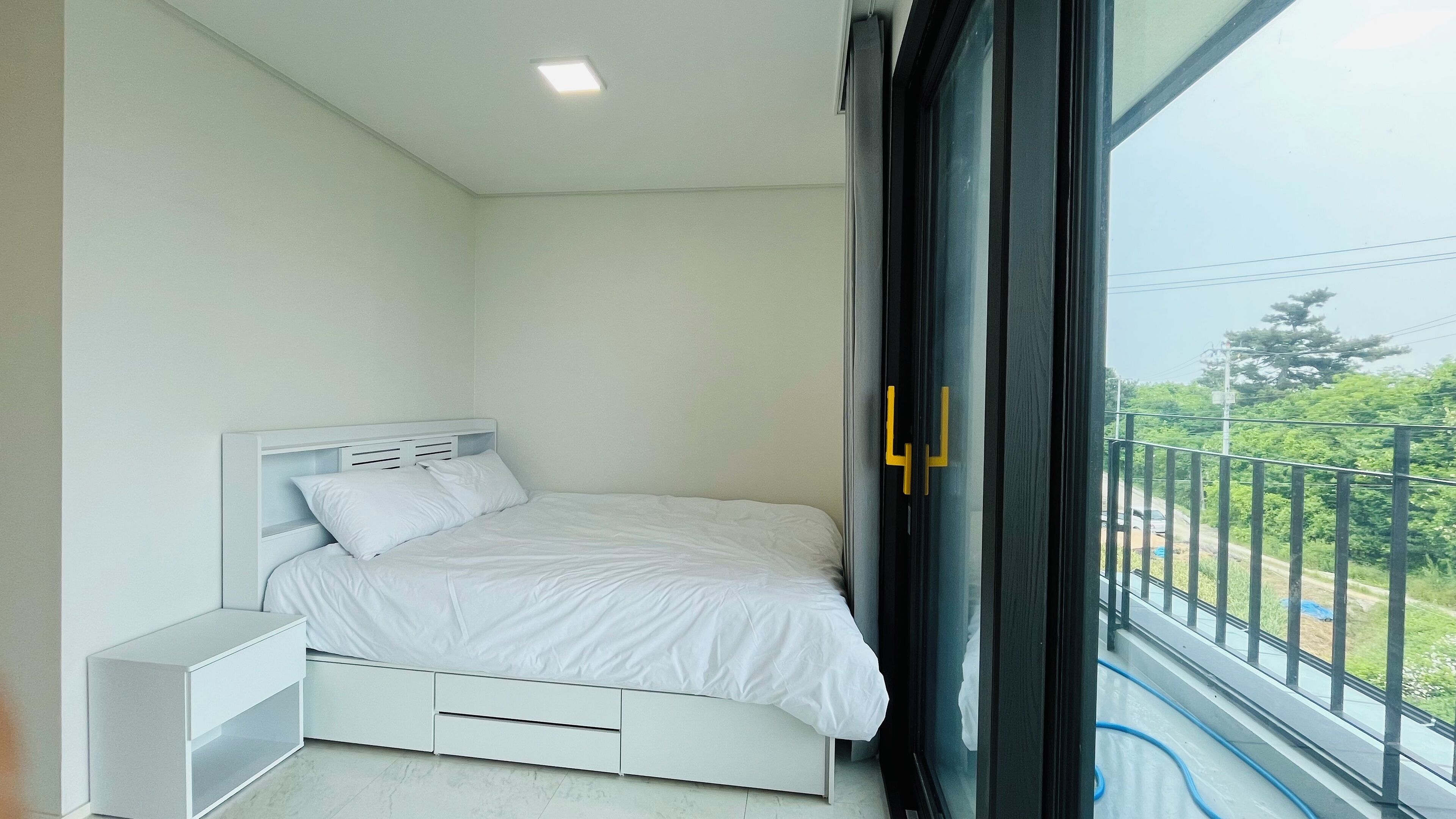 Quarto com cama, mesa de cabeceira e uma grande janela com vista para varanda.