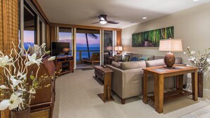 A spacious living room with a large window offering a view of the ocean, a ceiling fan, and a television. - Exquisite Oceanfront Beautifully Upgraded Corner Unit - Vrkcv (Kailua-Kona)