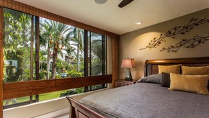 A bedroom with a large window, a bed with multiple pillows, a nightstand, and a wall-mounted lamp. - Exquisite Oceanfront Beautifully Upgraded Corner Unit - Vrkcv (Kailua-Kona)