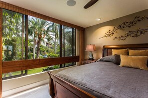 Ein Schlafzimmer mit großem Fenster, einem Bett mit vielen Kissen, einem Nachttisch und einer Wandlampe.
