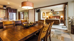 A spacious dining area with a wooden table and chairs, a kitchen with modern appliances, and a bedroom with a large bed and artwork on the wall. - Exquisite Oceanfront Beautifully Upgraded Corner Unit - Vrkcv (Kailua-Kona)