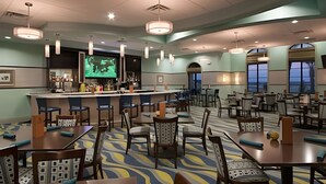 Un vestíbulo de hotel con una zona de bar, varios arreglos de asientos y una pantalla grande que muestra un programa.