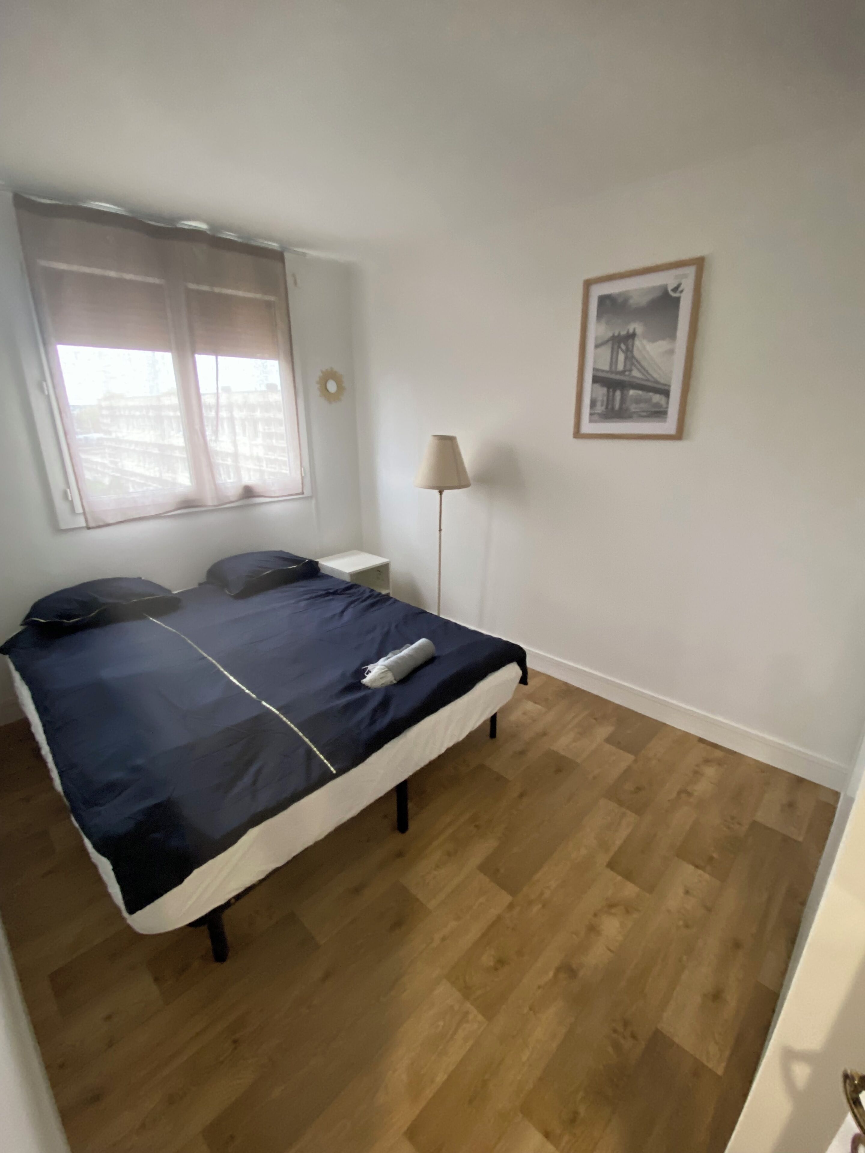Un dormitorio con cama, mesita de noche, lámpara, ventana con cortinas y un cuadro en la pared.