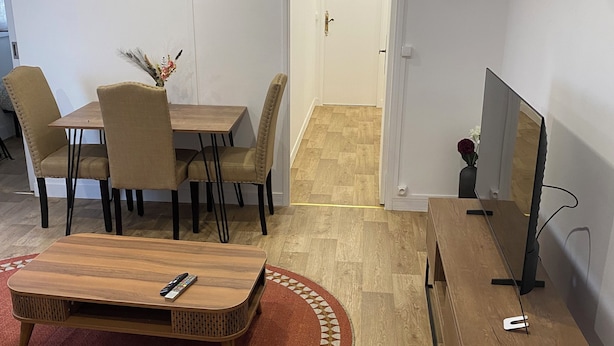 A modern living room with a wooden coffee table, beige sofa, and a TV stand. There is a dining area with a table and chairs, and a door leading to another room. - Pleasant apartment in the heart of the city (Orléans)