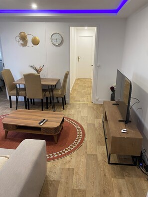 Una sala moderna con una mesa de centro de madera, un sofá color beige y un mueble para el televisor. Hay una zona de comedor con una mesa y sillas, y una puerta que da a otra habitación.