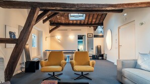 Un salon chaleureux avec un plafond haut, des poutres apparentes, un canapé, deux fauteuils, une table basse et une cheminée.