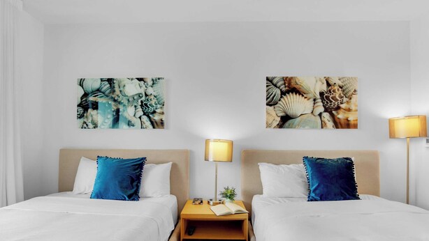 A hotel room with two beds, white bedding, blue pillows, a nightstand with a lamp, and two wall-mounted artworks. - Large 2-Bedroom Condo Near Ocean w Amenities 1009 (Hallandale Beach)