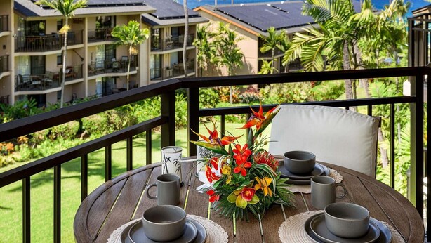 A balcony with a table set for two, overlooking a resort with multiple buildings and a view of the ocean. - Ocean View Top Floor Concierge Included! (Kailua-Kona)