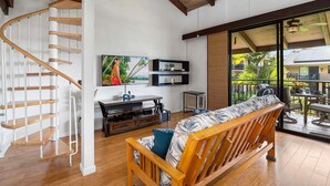A modern living room with a wooden staircase, a flat-screen TV, a sofa, and a balcony with outdoor furniture. - Ocean View Top Floor Concierge Included! (Kailua-Kona)