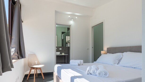 Une chambre d’hôtel avec un lit, une chaise, une salle de bain visible à travers une porte ouverte et une fenêtre avec des rideaux.