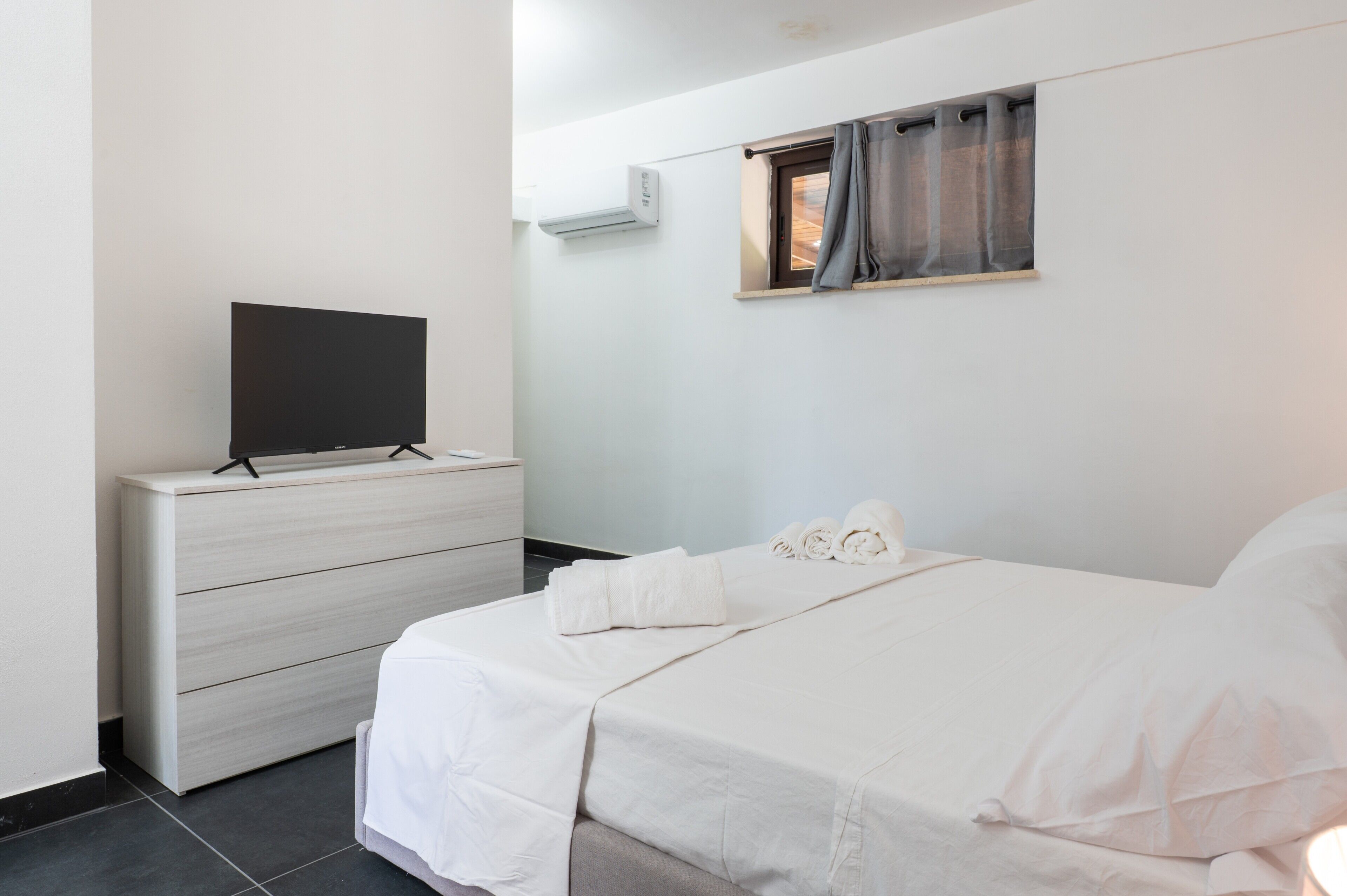 Une chambre à coucher avec un lit, un téléviseur posé sur une commode, une fenêtre avec des rideaux et un appareil de climatisation.
