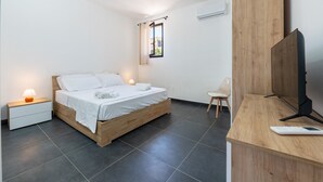A modern bedroom with a wooden bed, a bedside table with a lamp, a TV on a wooden stand, and a window with curtains. - Room 'Altavilla 3' with Shared Garden and Air Conditioning (Altavilla Milicia)