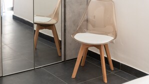 Een stoel met een transparante zitting en houten poten, geplaatst tegen een muur waar een spiegel de stoel in weerspiegelt.