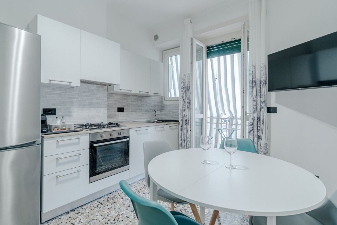 Cucina moderna con mobili bianchi, piano cottura e forno. C'è un tavolo da pranzo con sedie e un bicchiere da vino.