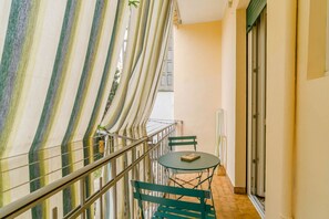 Un balcone con tende a righe, ringhiera in vetro, un tavolino con un libro e una sedia verde.