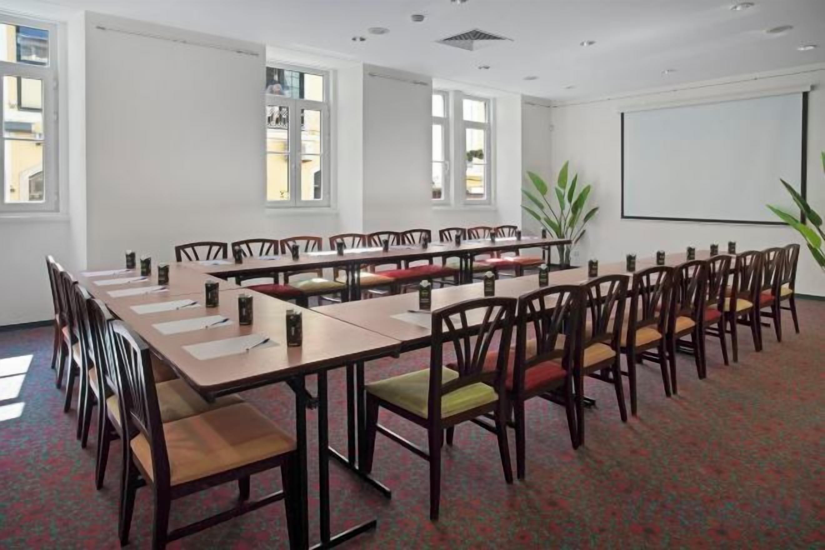 Un salón de conferencias con mesas largas y sillas, una pantalla de proyector y ventanas.