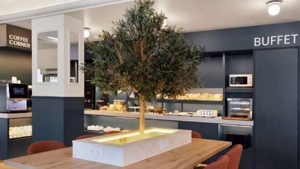 A modern dining area with a central table, chairs, a decorative tree, and a buffet counter in the background. - B&B HOTEL Figueira Da Foz (Figueira da Foz)